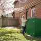 Homeowner using ToteBoxx residential bin enclosure in spring with blooming cherry blossoms, preparing property for spring wildlife season